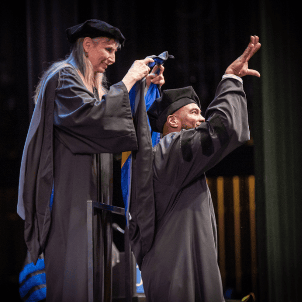 A person in academic regalia places a hood over a graduate’s shoulders on stage; the graduate waves while smiling, both wearing black gowns and caps.