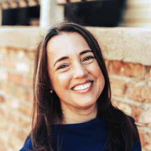 A woman with long brown hair and a blue top smiles outdoors in front of a brick wall.