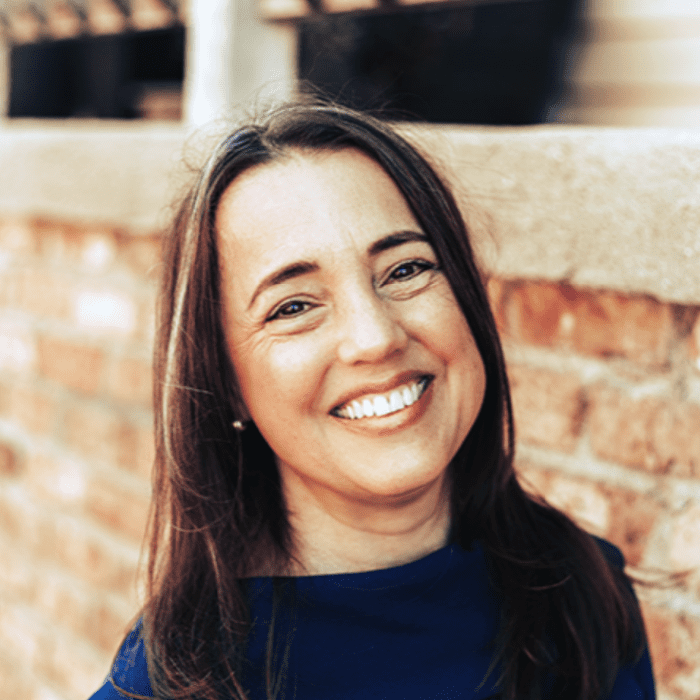 A woman with long brown hair and a blue top smiles outdoors in front of a brick wall.