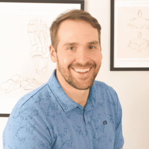 A man with short brown hair and a beard, wearing a blue polo shirt, smiles at the camera. Anatomical posters are visible on the wall behind him.