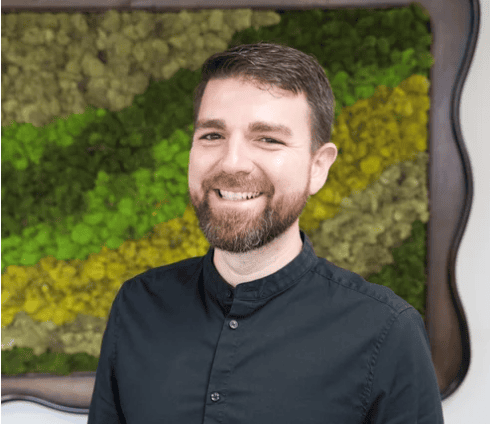 A man with short brown hair and a beard, wearing a dark button-up shirt, stands smiling in front of a green and yellow moss wall art.