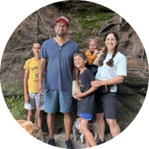 Five people and two dogs stand outdoors in front of a rocky, tree-covered area, smiling at the camera. Showcasing one of the 2024 PBPH Success Stories, some are holding rocks and everyone is dressed in casual clothes.
