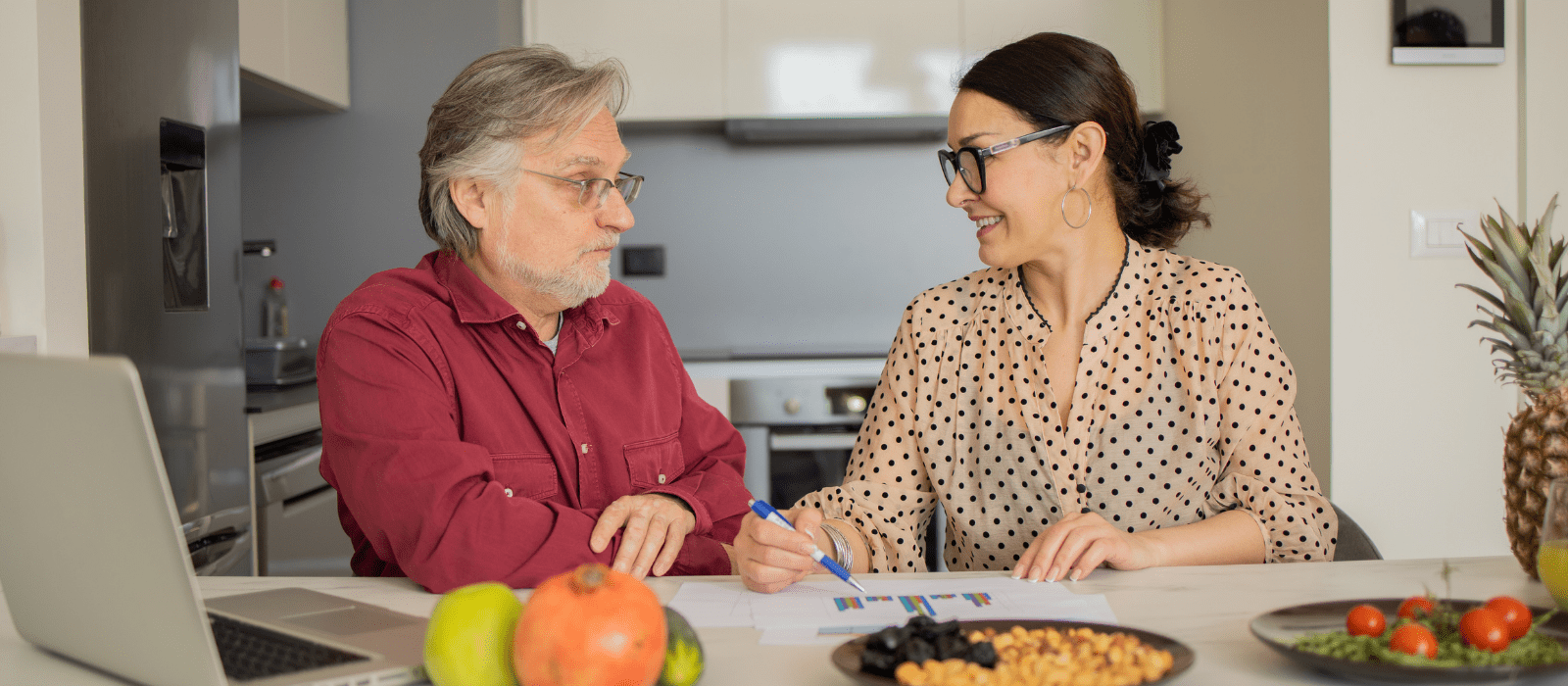 Two people sit at a kitchen table discussing documents. A laptop, fruits, and snacks are on the table. The woman is writing, and both appear engaged in conversation.