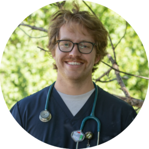 A person with glasses and a mustache, wearing navy scrubs and a stethoscope, stands outdoors in front of green foliage, exemplifying the dedication featured in 2024 PBPH Success Stories.